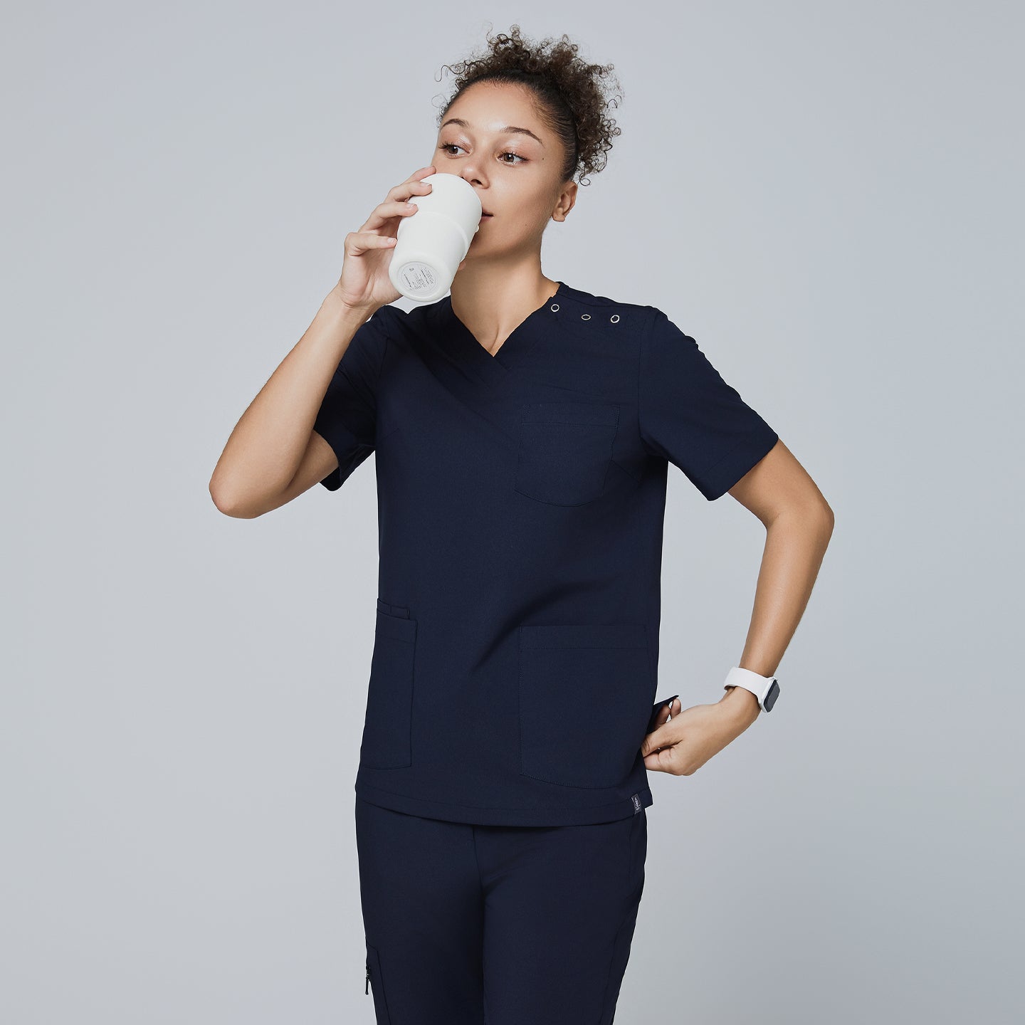 Image of Eco navy scrub top with shoulder buttons, v-neck, and two front pockets. Model holding tumbler and placing hand in pocket for a functional look,Eco Navy